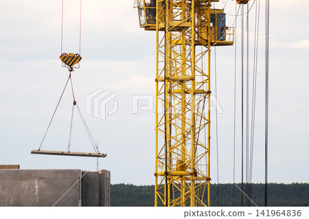 Heavy duty yellow tower crane with large hook lifting rectangular precast concrete panel for construction of multistory residential panel house, cloudy sky and distant forest line background Heavy duty yellow tower crane with large hook lifting rectangular precast concrete panel for construction of multistory residential panel house, cloudy sky and distant forest line background 141964836