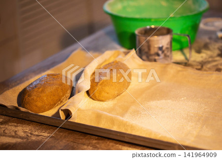 Freshly prepared bread dough loaves resting on baking sheet with flour dusting at bakery production facility, awaiting baking process Freshly prepared bread dough loaves resting on baking sheet with flour dusting at bakery production facility, awaiting baking process 141964909