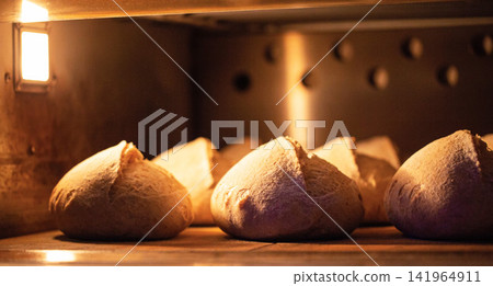 Warm loaves artisan bread baking inside industrial oven, golden light illuminating crust bread, traditional baked goods production Warm loaves artisan bread baking inside industrial oven, golden light illuminating crust bread, traditional baked goods production 141964911