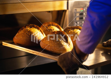 Baker hands placing warm, golden-crusted artisan bread loaves on metal peel into hot industrial oven, demonstrating manual production. 141964912
