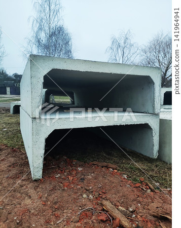 Two large precast concrete culvert sections stacked outdoors on construction site with bare trees and cloudy sky in background, showing material for infrastructure project. Two large precast concrete culvert sections stacked outdoors on construction site with bare trees and cloudy sky in background, showing material for infrastructure project. 141964941