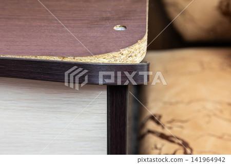 Close-up detail of edge of particle board furniture showing exposed chipboard core and peeling laminate surface, screw head visible, worn corner construction of cabinet lid or shelf Close-up detail of edge of particle board furniture showing exposed chipboard core and peeling laminate surface, screw head visible, worn corner construction of cabinet lid or shelf 141964942