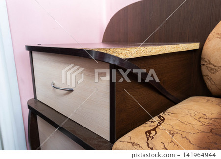 Damaged laminate surface of nightstand, bedside furniture with visible particle board and peeling edge, clearly showing need for urgent repair, typical bedroom interior setting. Damaged laminate surface of nightstand, bedside furniture with visible particle board and peeling edge, clearly showing need for urgent repair, typical bedroom interior setting. 141964944