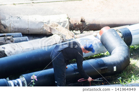 Worker performing repair work on large black heating pipes in outdoor setting during daytime, bent over pipe connection with tools and cable in hands, showing process of infrastructure maintenance. Worker performing repair work on large black heating pipes in outdoor setting during daytime, bent over pipe connection with tools and cable in hands, showing process of infrastructure maintenance. 141964949