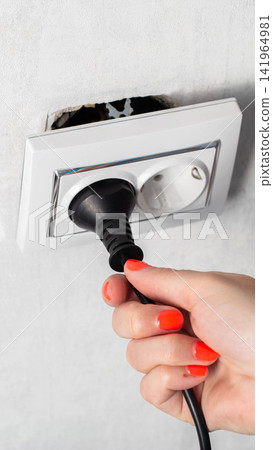 Person hand with vibrant orange nail polish inserting black power plug into white electrical outlet on rough textured wall, showing exposed wiring behind faulty socket, vertical photo Person hand with vibrant orange nail polish inserting black power plug into white electrical outlet on rough textured wall, showing exposed wiring behind faulty socket, vertical photo 141964981
