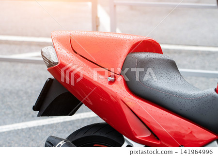 Wet red sport motorcycle rear section detail showing water droplets on bodywork and black seat, tail light, exhaust, and part of rear wheel in outdoor parking lot with sunlight flare. Wet red sport motorcycle rear section detail showing water droplets on bodywork and black seat, tail light, exhaust, and part of rear wheel in outdoor parking lot with sunlight flare. 141964996