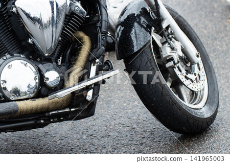 Close-up of motorcycle front section displaying chrome engine components, wrapped exhaust pipe, black wet fender, disc brake on front wheel, parked on damp asphalt road Close-up of motorcycle front section displaying chrome engine components, wrapped exhaust pipe, black wet fender, disc brake on front wheel, parked on damp asphalt road 141965003