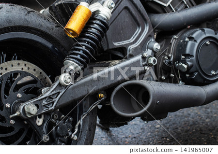 Detail shot of rear section of modern black motorcycle showing wet tire, shiny brake disc, golden suspension shock absorber, and matte black exhaust pipe with raindrops, parked outdoors on wet asphalt Detail shot of rear section of modern black motorcycle showing wet tire, shiny brake disc, golden suspension shock absorber, and matte black exhaust pipe with raindrops, parked outdoors on wet asphalt 141965007