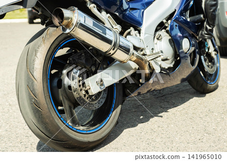 Close up view rear wheel sport motorcycle parked outdoors on asphalt during sunny day. Focus on powerful machine detail, including shiny chrome exhaust pipe, large black tire, and intricate disc brake Close up view rear wheel sport motorcycle parked outdoors on asphalt during sunny day. Focus on powerful machine detail, including shiny chrome exhaust pipe, large black tire, and intricate disc brake 141965010