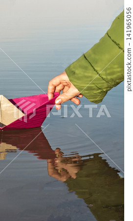 Human hand wearing green jacket gently placing homemade pink paper boat into calm water, reflections visible, simple childhood activity vertical photo Human hand wearing green jacket gently placing homemade pink paper boat into calm water, reflections visible, simple childhood activity vertical photo 141965056