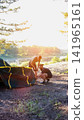 Two adult men assembling camping tent by serene lake water with lush green trees at golden hour sunset light, preparing for outdoor adventure, vertical photo 141965161