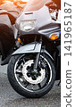 Front view of parked sport motorcycle with grey and black body, showing wheel, brake disc, and suspension after rain, drops visible, sunlight reflection on upper part. 141965187