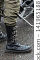 Close up view of person leg with heavy black combat boot featuring multiple silver spikes and buckles, olive green cargo pants, wet asphalt surface, motorcycle footpeg visible, vertical photo format 141965188