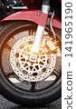 Close-up detail front wheel red motorcycle, wet fender and tire with raindrops after rain, disc brake system visible, sun flare on metal components, modern vehicle standing on asphalt ground 141965190