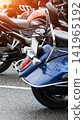 Rear view of parked blue cruiser motorcycle with chrome details and red taillight wet from rain or water droplets, passenger backrest and license plate frame visible, blurred motorcycles in background 141965192