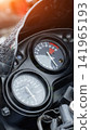 Close-up detail of motorcycle dashboard with speedometer showing speed in kilometers per hour and tachometer displaying engine RPM, raindrops visible on windshield, bright lens flare top left 141965193