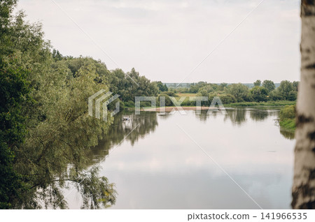 Wide scenic river with quiet current and dense wooded banks under a gloomy overcast sky, peaceful nature landscape Wide scenic river with quiet current and dense wooded banks under a gloomy overcast sky, peaceful nature landscape 141966535
