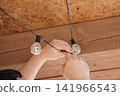Close-up of human hands hanging a warm string of decorative light bulbs from the ceiling, home interior decoration concept 141966543