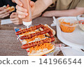 Hand pouring rich savory sauce over a grilled sausage in a hot dog topped with Korean-style spicy carrots, street food 141966544