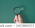 Hand holding a classic magnifying glass against a solid green background with copy space, ecological research concept 141966553
