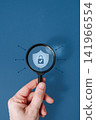 Hand holding magnifying glass over protective shield icon on blue background, data security and insurance concept 141966554