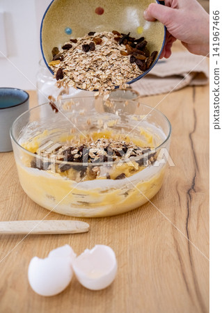 Homemade breakfast process. Healthy recipe. Female chef hand pouring oat flakes raisins in dough bowl on kitchen counter with eggshell. Homemade breakfast process. Healthy recipe. Female chef hand pouring oat flakes raisins in dough bowl on kitchen counter with eggshell. 141967466
