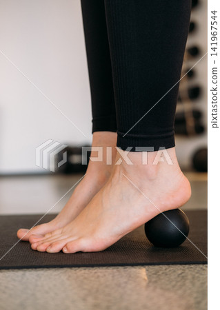 Foot massage. Rehabilitation exercise. Barefoot woman rolling black ball on mat for sole heel health in fitness studio. Physical therapy for leg. Foot massage. Rehabilitation exercise. Barefoot woman rolling black ball on mat for sole heel health in fitness studio. Physical therapy for leg. 141967544
