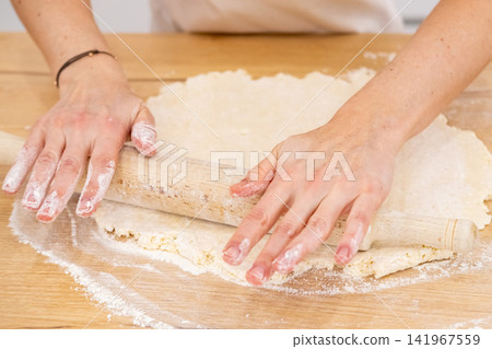 Baking process. Rolling dough pin. Female chef hand flattening sweet mass on home wooden kitchen table covered with flour making pastry. Baking process. Rolling dough pin. Female chef hand flattening sweet mass on home wooden kitchen table covered with flour making pastry. 141967559