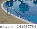 Stainless steel pool ladder leading into clear blue swimming pool 141967734