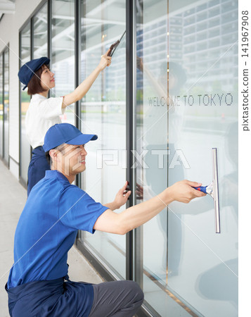 Two building maintenance staff members, a man and a woman, wiping windows. Two building maintenance staff members, a man and a woman, wiping windows. 141967908