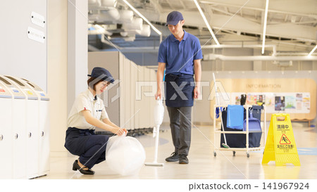 A woman gathering garbage bags and a male cleaning staff member with a vacuum cleaner. A woman gathering garbage bags and a male cleaning staff member with a vacuum cleaner. 141967924
