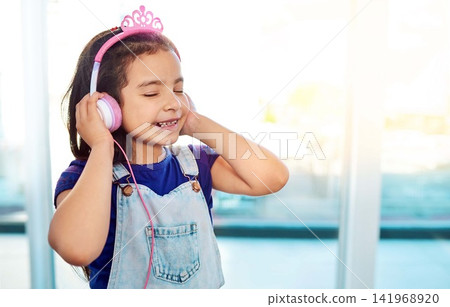 Young child, headphones and music listening in house with streaming song, radio sound and girl podcast. Happiness, kid and earphones for online media with smile, fun audio and playlist by home window Young child, headphones and music listening in house with streaming song, radio sound and girl podcast. Happiness, kid and earphones for online media with smile, fun audio and playlist by home window 141968920