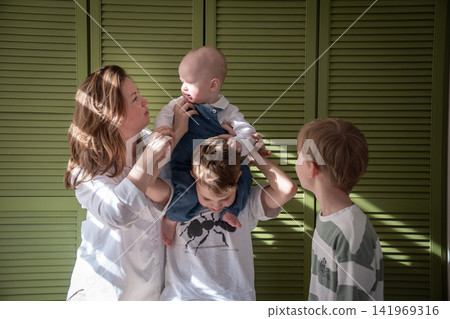 Mother poses with three sons while baby sits on older brothers shoulders during playful family moment at home. Motherhood, brotherhood, sibling bond, parenting, childhood, family connection 141969316