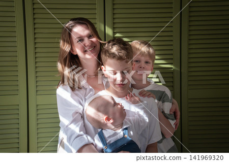Mother embraces three sons while baby sits on older brothers lap during close family moment at home. Motherhood, brotherhood, sibling bond, parenting, childhood, family love, caregiving, togetherness Mother embraces three sons while baby sits on older brothers lap during close family moment at home. Motherhood, brotherhood, sibling bond, parenting, childhood, family love, caregiving, togetherness 141969320