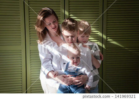 Mother holds baby while two older brothers gather closely around younger sibling during family moment at home. Motherhood, brotherhood, sibling care, parenting, childhood, family bond, affection Mother holds baby while two older brothers gather closely around younger sibling during family moment at home. Motherhood, brotherhood, sibling care, parenting, childhood, family bond, affection 141969321