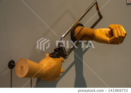 Wooden mannequin arms with hacksaw creating surreal self cutting wall installation 141969733