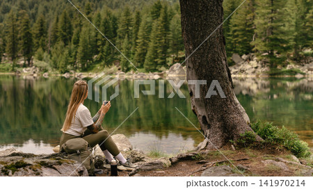 Woman Hiker with Backpack Sitting by Mountain Lake Woman Hiker with Backpack Sitting by Mountain Lake 141970214