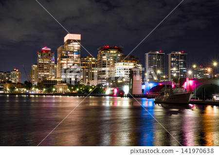Coastal City Buildings Glow Above Calm Nighttime Water Coastal City Buildings Glow Above Calm Nighttime Water 141970638