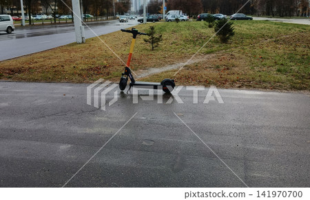 Electric scooter parked on damp sidewalk by an autumn park strip beside a busy multi-lane street, wet pavement and passing cars highlighting urban micromobility life Electric scooter parked on damp sidewalk by an autumn park strip beside a busy multi-lane street, wet pavement and passing cars highlighting urban micromobility life 141970700