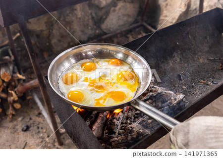 View to cooking eggs in a stainless steel pan over an open fire outdoors View to cooking eggs in a stainless steel pan over an open fire outdoors 141971365
