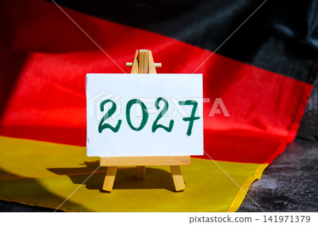Paper note with text 2027 on National symbol of Germany. German wave silky flag background. Financial outlook planning, new year annual report Paper note with text 2027 on National symbol of Germany. German wave silky flag background. Financial outlook planning, new year annual report 141971379