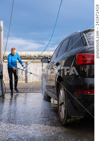 Man Washing Car at Self Service Car Wash. Applying thick foam with a pressure washer. The scene takes place in an outdoor washing bay with modern self-service equipment, automotive accessories Man Washing Car at Self Service Car Wash. Applying thick foam with a pressure washer. The scene takes place in an outdoor washing bay with modern self-service equipment, automotive accessories 141971419