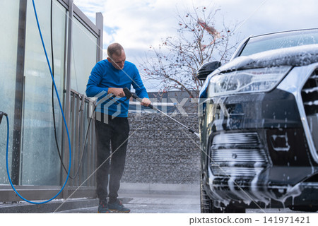 Man cleans his vehicle at self service car wash station using professional washing equipment. Rinsing the vehicle with high-pressure water stream system. Outdoor washing bay Man cleans his vehicle at self service car wash station using professional washing equipment. Rinsing the vehicle with high-pressure water stream system. Outdoor washing bay 141971421