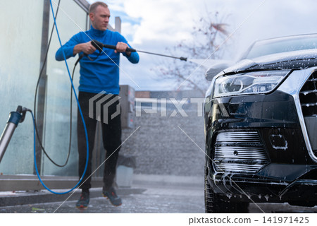 Man cleans his vehicle at self service car wash station using professional washing equipment. Rinsing the vehicle with high-pressure water stream system. Outdoor washing bay 141971425
