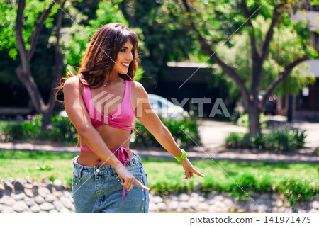 Beautiful mature Latina Arab brunette dancing in an outdoor park. Wearing retro pink clothing and jeans, with makeup, she dances with style, attitude, beauty, and freedom. Beautiful mature Latina Arab brunette dancing in an outdoor park. Wearing retro pink clothing and jeans, with makeup, she dances with style, attitude, beauty, and freedom. 141971475