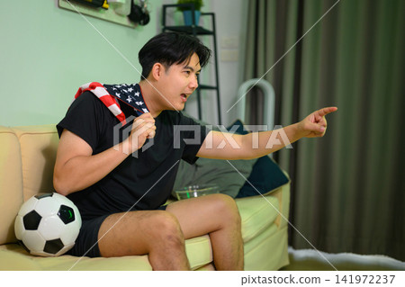 Excited Man Cheering Football Game at Home with American Flag and Soccer Ball on Sofa 141972237