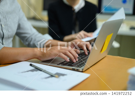 Business colleagues collaborating at desk with laptop, documents and sticky note in modern office workspace Business colleagues collaborating at desk with laptop, documents and sticky note in modern office workspace 141972252