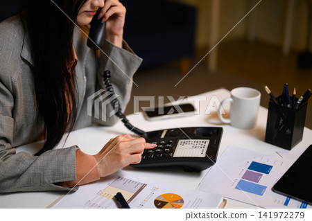 Businesswoman Talking on Landline Phone at Office Desk with Financial Charts and Documents Businesswoman Talking on Landline Phone at Office Desk with Financial Charts and Documents 141972279