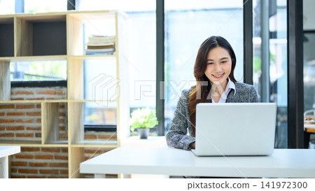 Smiling Young Asian Businesswoman Working on Laptop at Modern Office Desk with Brick Wall Background Smiling Young Asian Businesswoman Working on Laptop at Modern Office Desk with Brick Wall Background 141972300