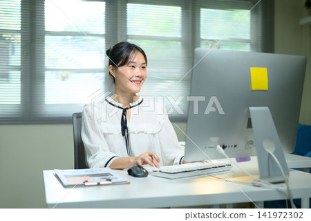 Smiling Young Asian Businesswoman Working at Computer Desk in Modern Office Smiling Young Asian Businesswoman Working at Computer Desk in Modern Office 141972302